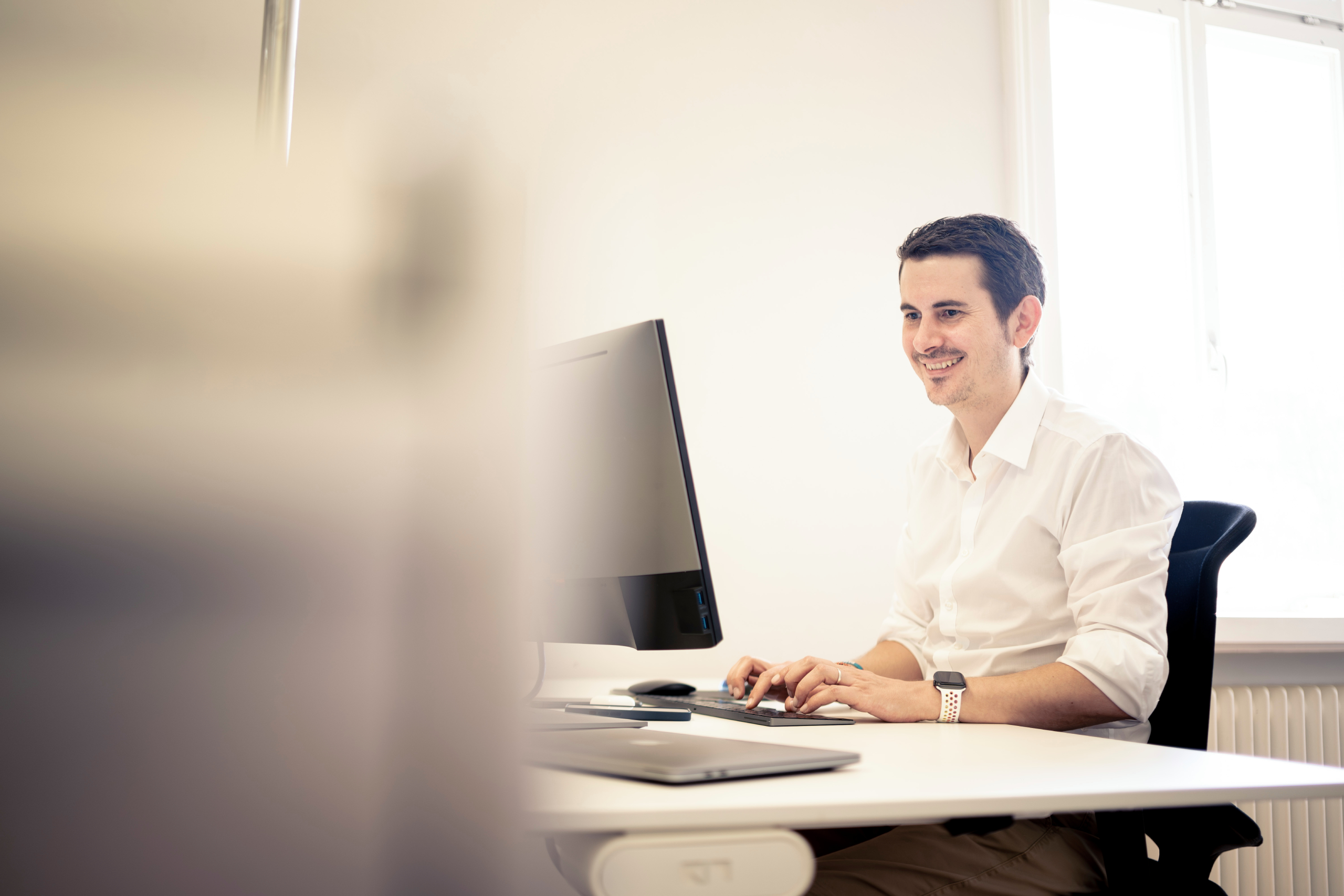 Mann mit weissem Hemd der in einem Büro am Computer sitzt und lacht. 