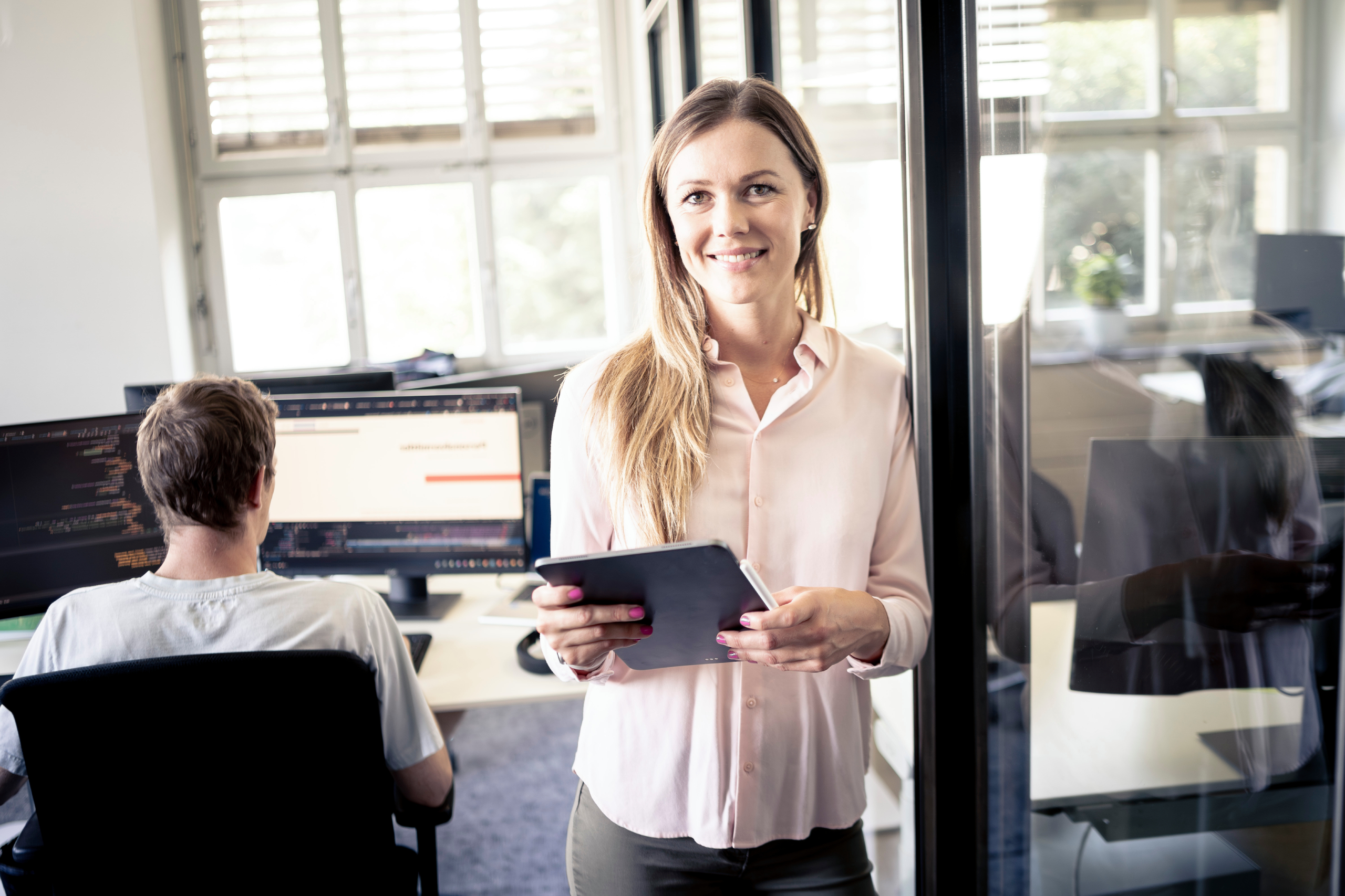 Eine Frau die mit Tablet in einem Büro steht und sich an einer Glaswand anlehnt. Auf dem Tablet verwendet sie das Recruiting-Tool Jobbooster. 