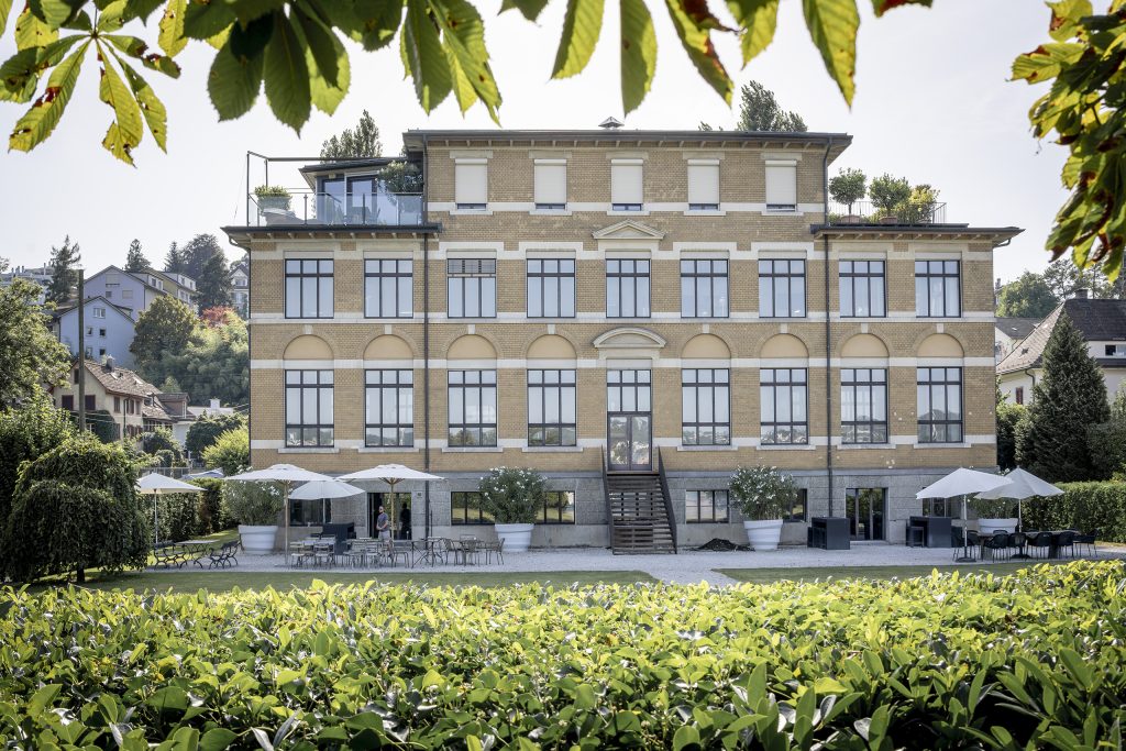 Eine grosse Villa mit vielen Fenstern im Vordergrund ist eine grüne Wiese zu sehen.