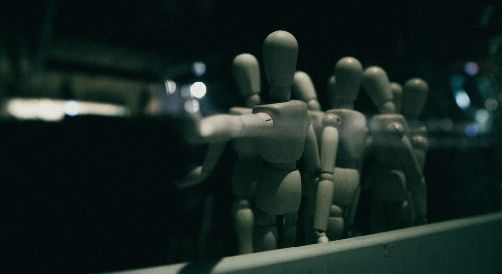 Eine Gruppe von hölzernen Zeichenpuppen steht in einer dunklen, unscharfen Umgebung. Symbolbild für Unconscious Bias.